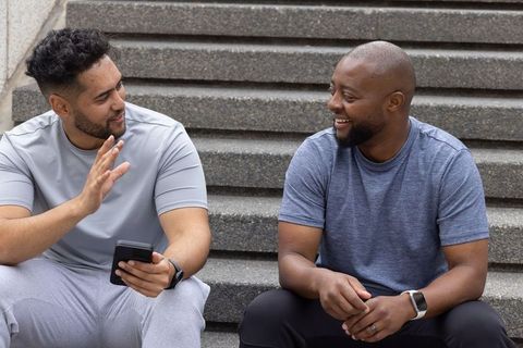 African american men chatting on steps wearing activewear and smartwatches, using phone