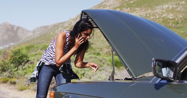 African american woman talking on smartphone while standing near her broken down car on road. road trip travel and adventure concept