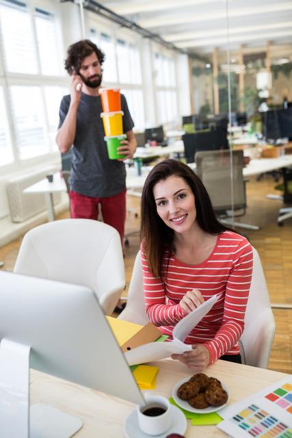 Female graphic designer in red striped shirt is analyzing a document at her desk, while a male colleague holds plastic containers in the background. The modern office space has an open, collaborative environment suitable for creative professionals. Great for advertisements or articles on modern workplace culture, team dynamics, office productivity, and creativity.