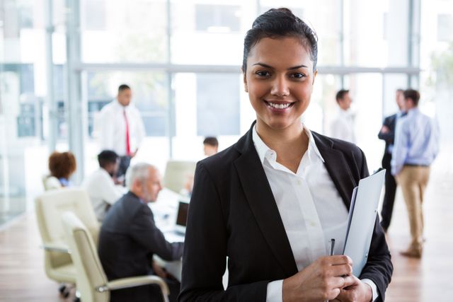 Businesswoman holding a folder, smiling at camera in a modern office environment. Team members are having a discussion in the background. Ideal for corporate websites, professional services promotions, leadership articles, and diversity in the workplace campaigns.