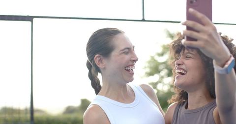 Diverse friends laughing while taking pink phone selfie outdoors in athleisure, sunlit happiness