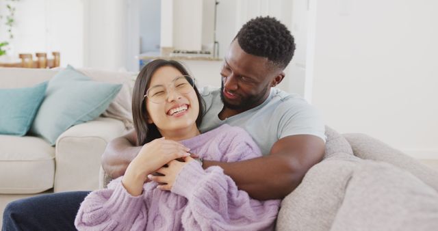 A warm and inviting scene of an interracial couple embracing on a sofa in a cozy living room. Ideal for use in advertisements, articles, or social media posts depicting love, happiness, and togetherness in home settings. Perfect for highlighting diversity and modern relationships.