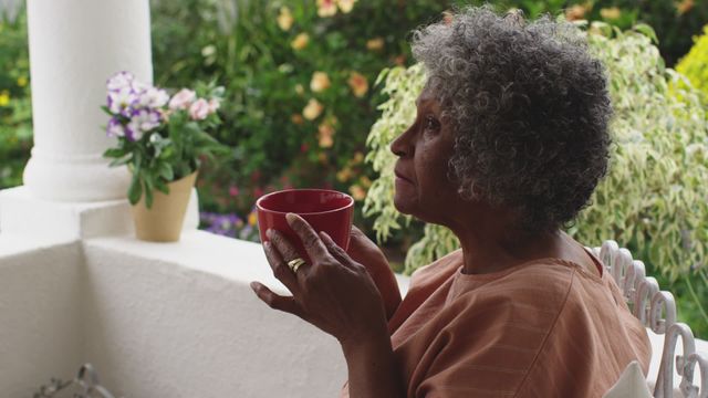 Senior African American woman enjoying a peaceful moment drinking coffee outdoors on the porch, exuding a sense of relaxation and contemplation. Could be used in materials focused on retirement lifestyle, pandemic social distancing life, serene moments, or elderly well-being.