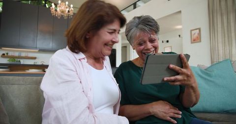 Senior women laughing while video-calling on tablet in cozy modern living room