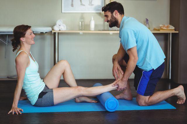 Physiotherapist assisting woman with leg therapy using foam roller in clinic. Ideal for illustrating physical therapy sessions, rehabilitation exercises, healthcare services, and wellness programs. Useful for websites, brochures, and articles related to physiotherapy, sports injury recovery, and fitness training.