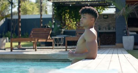 Young man relaxing on wooden pool deck beside turquoise water under sunlit pergola