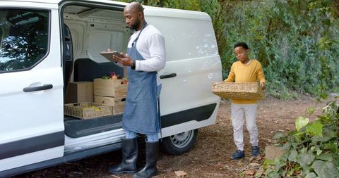 African american vendor and boy loading fresh produce into cargo van checking tablet