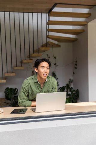 Asian man working remotely at modern counter with laptop, tablet and floating staircase