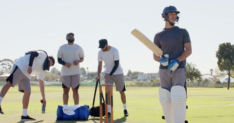 Male cricket teammates preparing on grassy pitch with bat, helmet, pads and kit bag