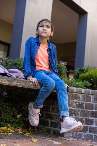 Asian girl sitting on bench in urban courtyard wearing denim jacket, coral tee, pink sneakers