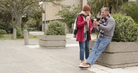 Mature couple enjoying coffee while sharing smartphone in urban plaza, scarves, fall day