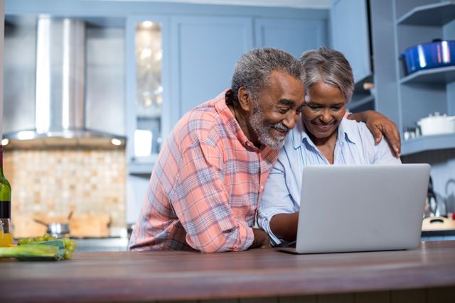 Elderly couple smiling while using a laptop in a modern kitchen. Ideal for themes related to senior lifestyle, technology use among older adults, home life, and family bonding. Can be used in advertisements, blogs, and articles focusing on aging, technology, and family relationships.