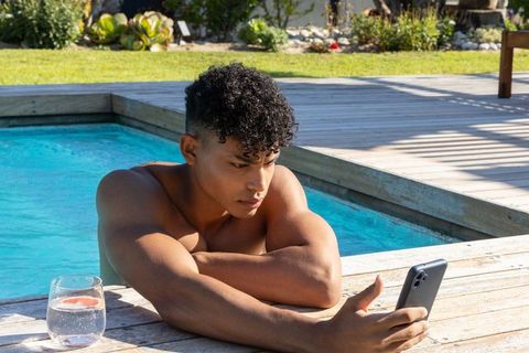 Young man relaxing poolside checking smartphone with citrus drink on sunlit deck by turquoise water