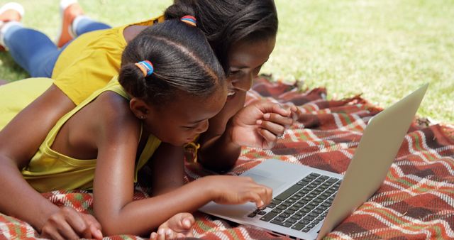 Mother and daughter lying on picnic blanket using laptop together outdoors. Suitable for themes involving family bonding, education, summer activities, technology usage in everyday life, parenting, and leisure.