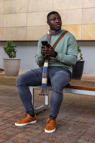 African american man sitting on urban bench wearing striped scarf and using smartphone