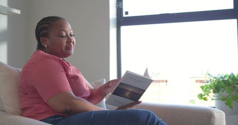 African american woman reading brochure on sunlit sofa near window, minimalist living room