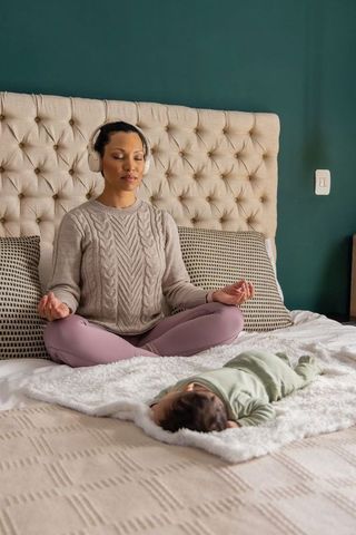 Calm mother meditating on bed with newborn, headphones and cozy bedroom retreat