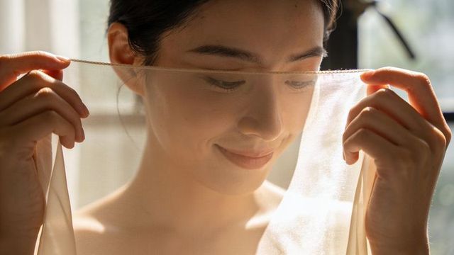 Asian woman holding sheer veil smiling softly backlit by window warm natural light