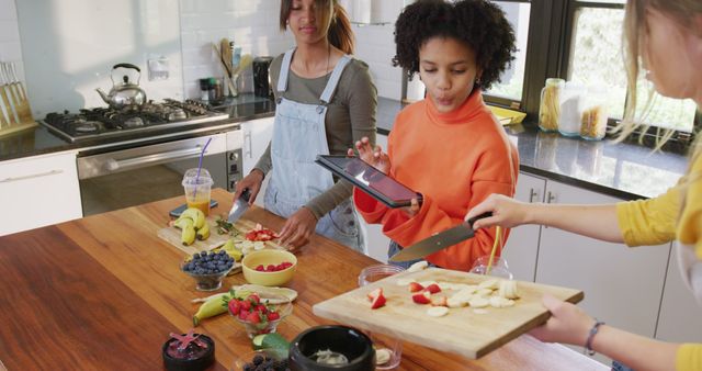 Group of children preparing a variety of fresh fruits while one holds a tablet for recipes in sunny modern kitchen. Useful for topics on healthy eating, cooking classes, child education, family activities, and DIY nutritious recipes.
