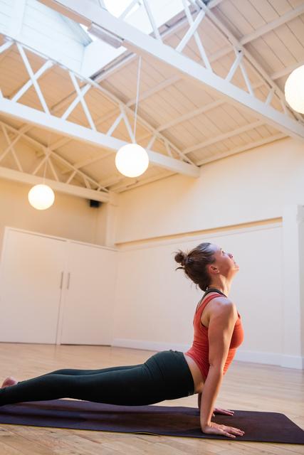 A woman is doing the cobra pose on an exercise mat in a bright, spacious fitness studio. This image is perfect for promoting yoga classes, wellness programs, fitness blogs, or health-related products. The serene environment and natural lighting highlight the calm and focused nature of yoga practice.