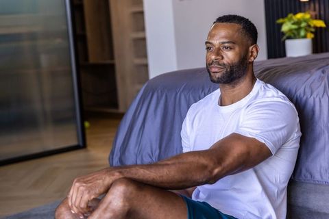 African american man sitting on bedroom floor relaxing and reflecting in minimalist interior