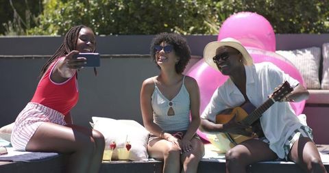 African american friends taking selfie at poolside while playing guitar and relaxing