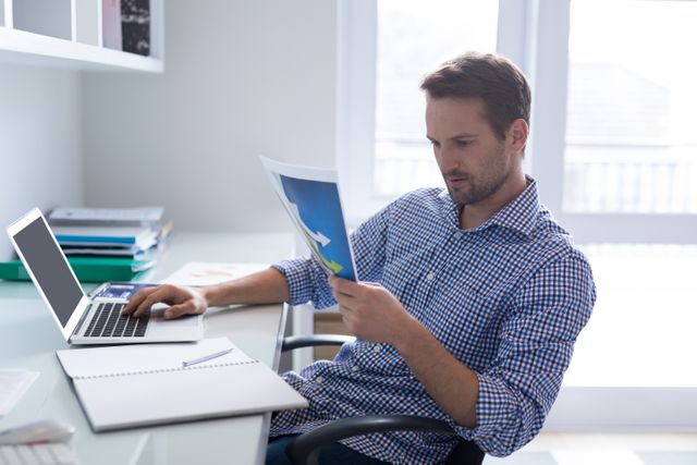 Man in checkered shirt analyzing a graph while using a laptop at a desk in a modern office. Ideal for use in business, finance, data analysis, and technology-related content. Perfect for illustrating concepts of professional work, office environments, and data-driven decision making.