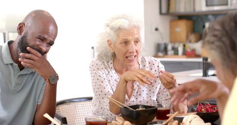 This scene captures a group of senior friends gathered around a table, enjoying a meal and conversation at home. Their expressions convey a sense of joy and camaraderie, making it an ideal image for promoting lifestyle concepts about senior living, friendship, and social gatherings. Utilize it in articles or advertisements focused on community—especially for seniors—or in content that highlights the joys of shared meals.