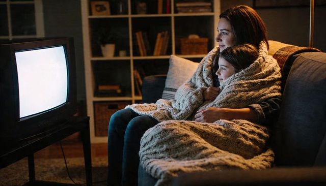 Mother and daughter snuggling under chunky knit blanket on couch watching tv during cozy evening