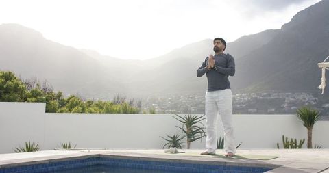 Indian man praying and meditating on rooftop yoga mat by pool with mountain valley view