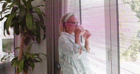 Senior woman sipping water by sliding glass door morning calm home relaxation natural light