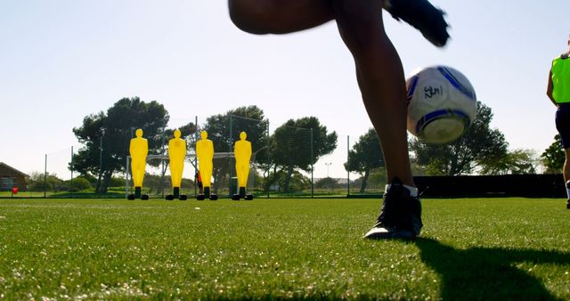 Close-up view of a soccer player practicing on an outdoor field, focusing on the motion of the kick. The background includes training mannequins and other players. This can be used for promoting sports training programs, soccer leagues, or athletic footwear.