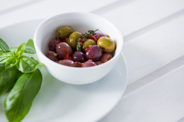 This image shows a close-up of marinated olives in a white bowl, accompanied by fresh basil leaves on a white plate. Ideal for use in culinary blogs, Mediterranean cuisine promotions, healthy eating articles, or gourmet food advertisements.
