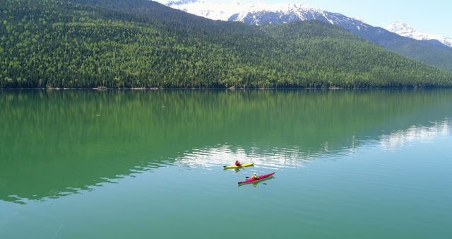 Two people are kayaking on a tranquil lake surrounded by lush green forests and snow-capped mountains. The vibrant colors of the kayaks contrast beautifully with the natural scenery, offering a sense of adventure and serenity.