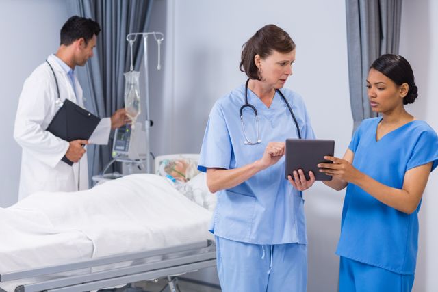 Medical team consisting of a doctor and nurse discussing patient care using a digital tablet in a hospital room. Another doctor is seen in the background attending to a patient in bed. Ideal for use in healthcare, medical technology, teamwork, and hospital-related content.