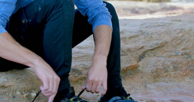 A man is lacing up his climbing shoes while seated on a rocky terrain during a hiking adventure. The image can be used for outdoor activity promotions, adventure blogs, climbing gear advertisements, fitness campaigns, or travel brochures focusing on nature and sports.