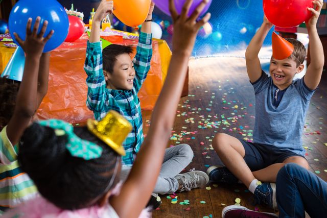 Children are sitting on the floor, raising balloons in the air, and smiling. They are surrounded by colorful confetti and party decorations. This image can be used for birthday party invitations, children's event promotions, or articles about kids' celebrations.