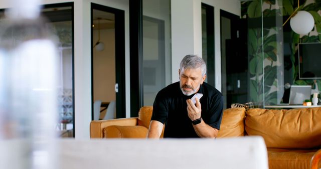 Senior man wearing black shirt, holding smartphone and engaged in a conversation. Seated in a contemporary living room featuring stylish decor and comfortable furniture. Perfect for depicting lifestyle, communication technology, and seniors connecting with loved ones.