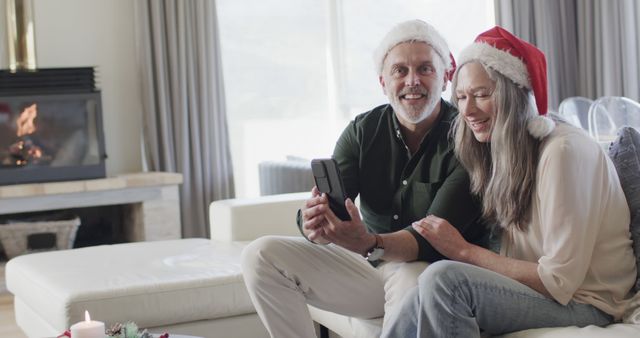 A couple wearing Santa hats enjoy a cheerful video call with family members while celebrating Christmas in their cozy living room. In the background, a warm fireplace creates a festive and inviting atmosphere. This can be used for holiday greeting cards, advertising festive tech gadgets, or promoting family-oriented services.