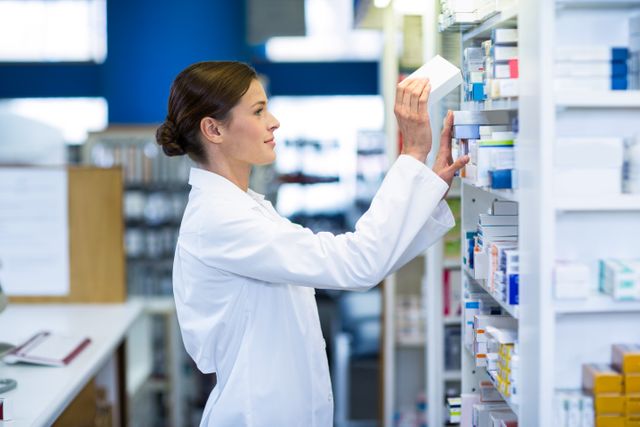 Pharmacist in white coat organizing medicine on shelves in a pharmacy. Ideal for use in healthcare, pharmaceutical, and medical industry content. Can be used in articles, advertisements, and educational materials related to pharmacy operations, medication management, and healthcare services.