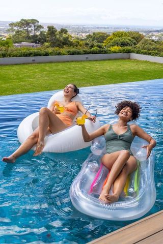 Laughing diverse women floating on pool inflatables enjoying summer drinks backyard relaxation