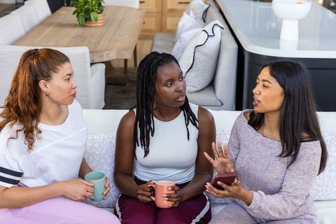 Three women chatting on sofa holding mugs and smartphone in cozy open-plan living room
