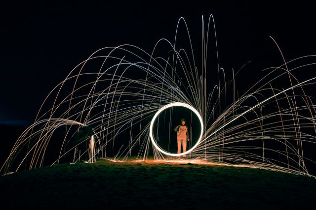 This captivating image shows a person standing inside a glowing circle at night, surrounded by dynamic light trails, creating a mesmerizing effect. The scene resembles a blend of light painting and performance art, captured with a long exposure to enhance the motion and impact of the light. Perfect for use in creative projects, artistic blogs, highlighting night photography techniques, or showcasing performance art in exotic locations.