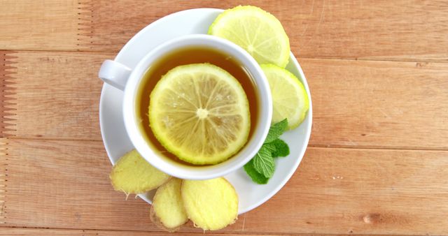 A cup of tea with a lemon slice floats on the surface, surrounded by fresh ginger and additional lemon slices on a wooden table, with copy space. The warm beverage setup suggests a home remedy approach, for wellness or soothing a cold.