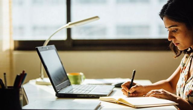 Sunlit home desk woman writing in notebook beside laptop focused on remote work
