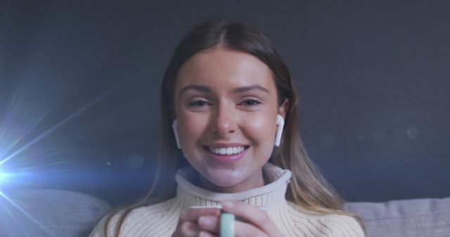 Young woman relaxing indoors, enjoying hot drink while smiling, wearing casual white sweater and airpods. Perfect for articles about relaxation, lifestyle, and indoor activities, warming up, coziness, happy moments.