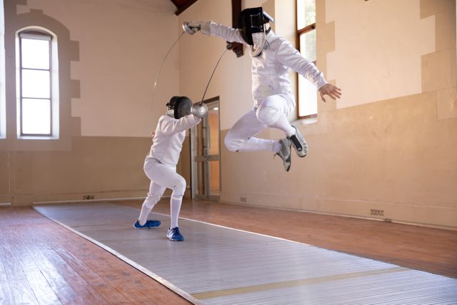 Two people in a fence duel, wearing protective fencing clothes, jumping in the air, in a gym. Sport and working out.
