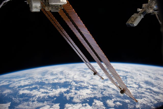 Image shows CubeSat deployment from the International Space Station using the Japanese robotic arm. Planet Labs Dove satellites visible against backdrop of Earth and space. Parts of ISS solar arrays in foreground. Useful for articles on space exploration, satellite technology, robotic arms in space operations, and Earth observation.