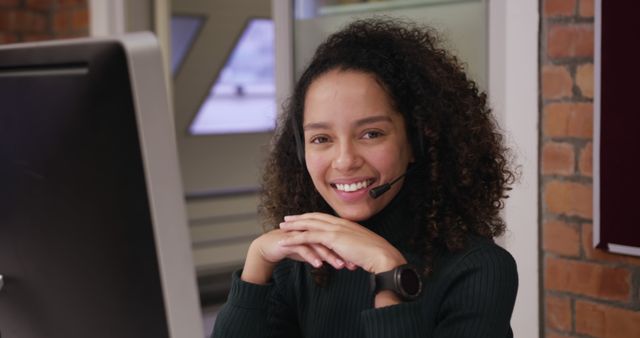 Young biracial woman wearing headset, working at computer. She has curly brown hair, light brown skin, and is wearing dark sweater