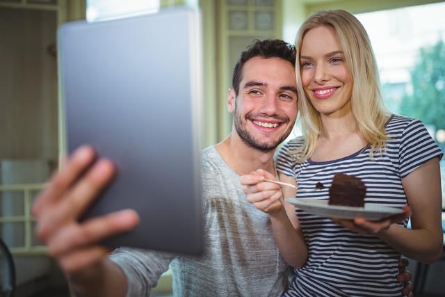 Smiling couple taking selfie from digital tablet while having pastries in cafÃ©